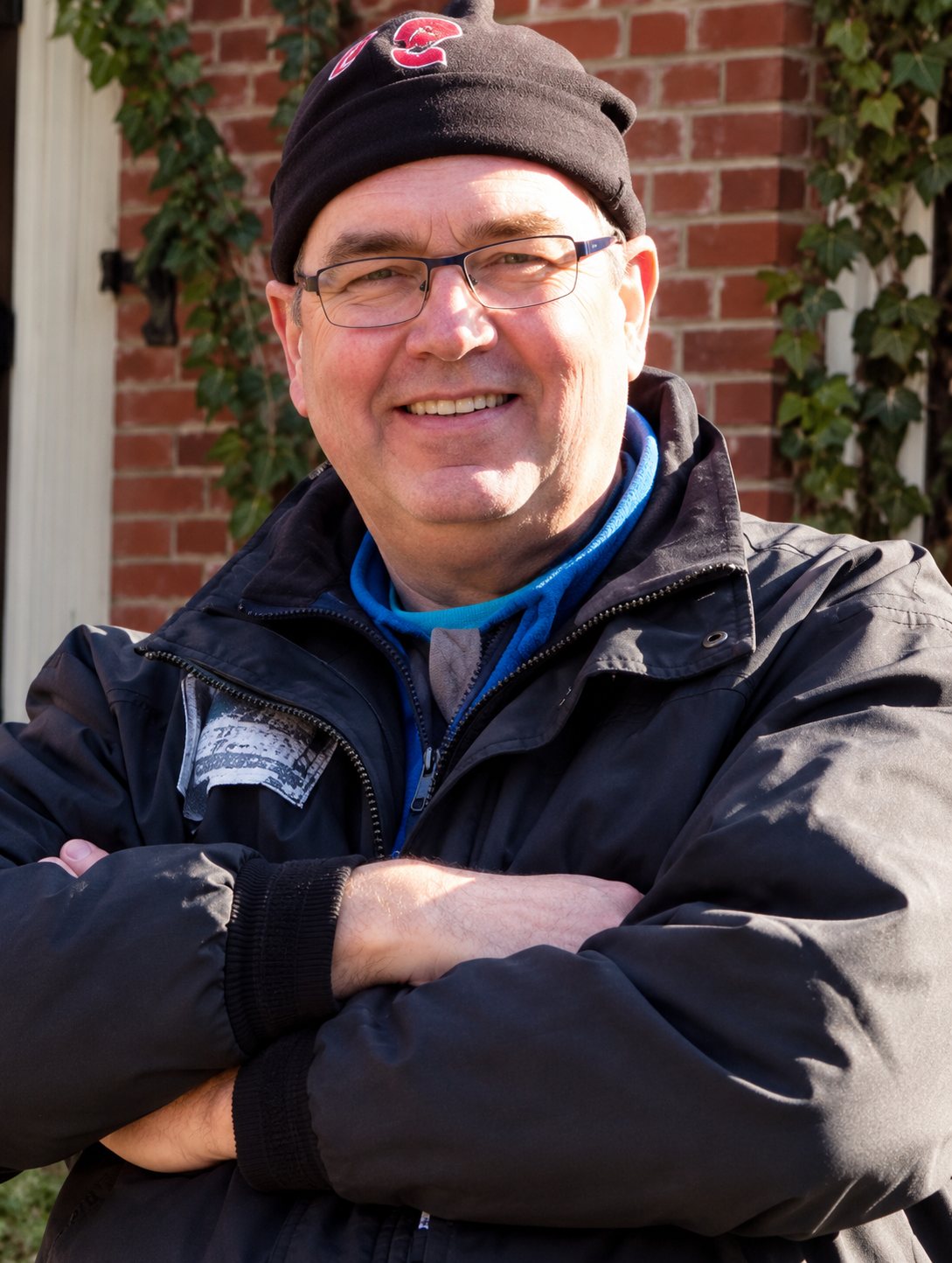 Homme souriant avec bonnet et lunettes, bras croisés devant un mur de briques.